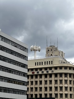 Several buildings with industrial and commercial designs stand under an overcast sky. An architectural structure features numerous antennas and communication devices on its rooftop.