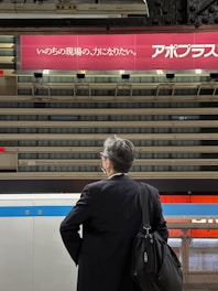 A man wearing a dark suit and carrying a shoulder bag stands on a train platform. He is looking towards the tracks, with a large advertisement featuring Japanese text visible in the background. The platform has a industrial, metallic design, and the lighting suggests an indoor setting.