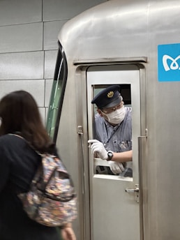 A train conductor wearing a uniform, face mask, and gloves peers from the window of a train door. A person with a colorful backpack passes by, partially out of frame. The metallic surface of the train and a blue sign with a white logo are visible.