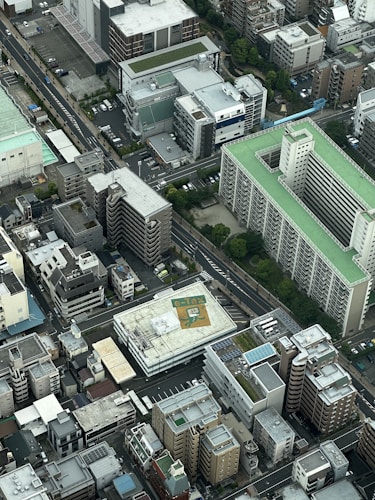 The aerial view captures a densely packed urban area with various multi-story buildings. The architecture includes both residential and commercial structures with flat rooftops. Some rooftops have solar panels, and one prominently features an 'e-tax' logo. The scene is dominated by concrete and asphalt but is interspersed with patches of greenery.