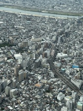 A sprawling urban landscape featuring a dense array of high-rise buildings and residential structures. A river runs horizontally across the background, bordered by green spaces. Roads and railway tracks intersect the cityscape, creating a network of transportation routes.
