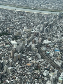 A sprawling urban landscape featuring a dense array of high-rise buildings and residential structures. A river runs horizontally across the background, bordered by green spaces. Roads and railway tracks intersect the cityscape, creating a network of transportation routes.