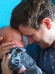 A gentle moment between a man and a newborn baby, with their foreheads touching. The man is cradling the baby tenderly, eyes closed in a peaceful expression. The baby is dressed in a soft, patterned outfit, asleep in the comfort of the man's arms.