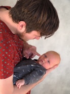 A person with brown hair is gently holding a baby, who has light hair and is wearing a dark onesie with words like 'happy' and 'milk' printed on it. The person is touching the baby's cheek with their finger, creating a tender and intimate moment. The background is a neutral gray texture.