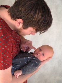 A person with brown hair is gently holding a baby, who has light hair and is wearing a dark onesie with words like 'happy' and 'milk' printed on it. The person is touching the baby's cheek with their finger, creating a tender and intimate moment. The background is a neutral gray texture.