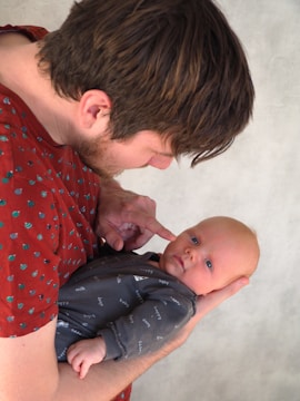 A person with brown hair is gently holding a baby, who has light hair and is wearing a dark onesie with words like 'happy' and 'milk' printed on it. The person is touching the baby's cheek with their finger, creating a tender and intimate moment. The background is a neutral gray texture.