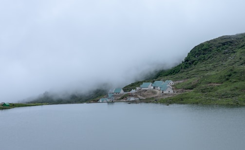 A serene mountain landscape with a cluster of buildings featuring green roofs nestled at the foot of a lush hillside. The foreground shows a calm body of water reflecting the overcast sky, with low-lying clouds partially obscuring the slopes.