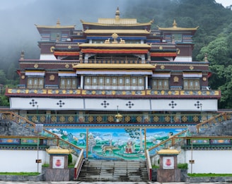 An ornate Buddhist monastery with intricate architectural details, adorned with gold accents and traditional motifs. The structure is surrounded by lush greenery and misty hills, creating an atmospheric backdrop. The monastery features a mural depicting serene landscapes and spiritual figures.