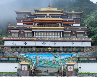 An ornate Buddhist monastery with intricate architectural details, adorned with gold accents and traditional motifs. The structure is surrounded by lush greenery and misty hills, creating an atmospheric backdrop. The monastery features a mural depicting serene landscapes and spiritual figures.