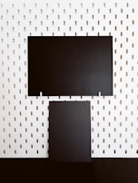 Two rectangular black panels are mounted on a white perforated board. The board features evenly spaced vertical oval holes, creating a regular pattern. The black panels contrast starkly against the white background.