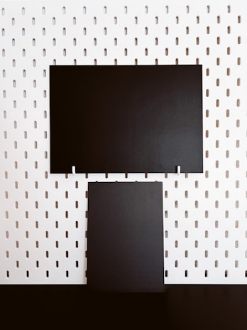 Two rectangular black panels are mounted on a white perforated board. The board features evenly spaced vertical oval holes, creating a regular pattern. The black panels contrast starkly against the white background.