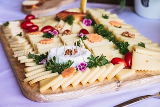 A wooden board is arranged with a variety of cheese slices, garnished with parsley and edible flowers. Cherry tomatoes and walnuts add color and texture. A wheel of soft cheese is centered, with toothpicks for serving. Delicate carving details on some cheeses and decorative slices of apple add sophistication.