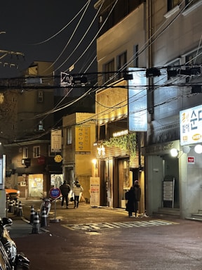 A nighttime urban scene features dimly lit streets with several buildings adorned with illuminated signs in Korean. Overhead, numerous power lines crisscross the street. A few pedestrians walk along the sidewalk, passing by a bicycle and traffic cones. A warm glow emanates from a storefront entrance, casting light on the surroundings.