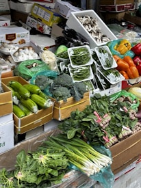 A vibrant display of various fresh vegetables neatly arranged in cardboard boxes and plastic packaging. The assortment includes green beans, spinach, broccoli, zucchini, bell peppers, mushrooms, and spring onions, creating a lively and colorful scene.