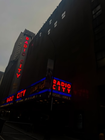 A well-known music hall with prominent neon signs displaying 'Radio City' in red and blue against a tall, dark building facade. The building has a textured exterior with small windows, and the sky appears overcast.