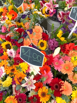 A vibrant display of mixed flower bouquets featuring an array of colors such as red, orange, pink, yellow, and purple. Each bouquet includes different types of flowers like gerberas, lilies, and sunflowers. Chalkboard signs with prices are visible, indicating a market setting.