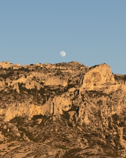A rugged and arid mountainous landscape is illuminated by warm evening light. The rocky terrain is dotted with sparse vegetation, and the moon is visible in the clear blue sky above.