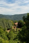 A rustic stone house with a red-tiled roof is nestled among lush green forests and rolling hills under a partly cloudy sky. The landscape is serene and abundant with verdant trees, creating a peaceful and secluded atmosphere.