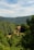 A rustic stone house with a red-tiled roof is nestled among lush green forests and rolling hills under a partly cloudy sky. The landscape is serene and abundant with verdant trees, creating a peaceful and secluded atmosphere.