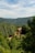 A rustic stone house with a red-tiled roof is nestled among lush green forests and rolling hills under a partly cloudy sky. The landscape is serene and abundant with verdant trees, creating a peaceful and secluded atmosphere.
