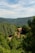A rustic stone house with a red-tiled roof is nestled among lush green forests and rolling hills under a partly cloudy sky. The landscape is serene and abundant with verdant trees, creating a peaceful and secluded atmosphere.