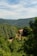 A rustic stone house with a red-tiled roof is nestled among lush green forests and rolling hills under a partly cloudy sky. The landscape is serene and abundant with verdant trees, creating a peaceful and secluded atmosphere.