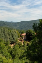 A rustic stone house with a red-tiled roof is nestled among lush green forests and rolling hills under a partly cloudy sky. The landscape is serene and abundant with verdant trees, creating a peaceful and secluded atmosphere.