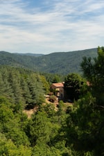A rustic stone house with a red-tiled roof is nestled among lush green forests and rolling hills under a partly cloudy sky. The landscape is serene and abundant with verdant trees, creating a peaceful and secluded atmosphere.