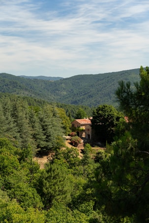 A rustic stone house with a red-tiled roof is nestled among lush green forests and rolling hills under a partly cloudy sky. The landscape is serene and abundant with verdant trees, creating a peaceful and secluded atmosphere.