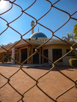 A mosque with a blue dome and a minaret is seen behind a chain-link fence. The building features intricate architectural details and is surrounded by a paved courtyard with some trees in the background.