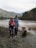 A family is standing near a river in a mountainous area. The woman is holding a small child, and a man stands beside them holding her hand. A brown dog sits nearby on the riverbank. The background features a river flowing gently with forested hills and mountains beyond.
