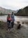 A family is standing near a river in a mountainous area. The woman is holding a small child, and a man stands beside them holding her hand. A brown dog sits nearby on the riverbank. The background features a river flowing gently with forested hills and mountains beyond.