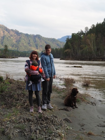 A family is standing near a river in a mountainous area. The woman is holding a small child, and a man stands beside them holding her hand. A brown dog sits nearby on the riverbank. The background features a river flowing gently with forested hills and mountains beyond.