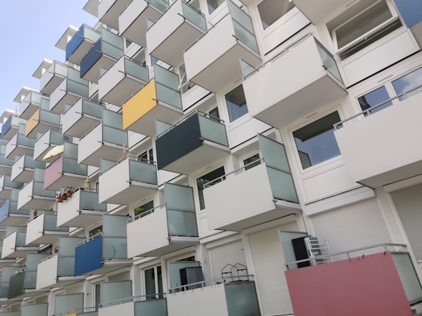 A modern apartment building with a unique geometric pattern of balconies, featuring various pastel-colored panels including yellow, blue, pink, and white. The balconies create a staggered, three-dimensional facade.