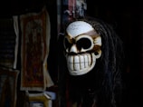 A skull-shaped mask with long black hair hangs against a dark background. The detailed carvings and sinister grin give it an eerie appearance. To the left, there are colorful, intricate fabric designs that appear to be tapestries or paintings.
