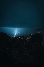 A dramatic lightning strike illuminates the night sky over a cityscape, with dark clouds above and some buildings in shadow. The scene captures the raw power and beauty of the storm.