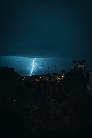 A dramatic lightning strike illuminates the night sky over a cityscape, with dark clouds above and some buildings in shadow. The scene captures the raw power and beauty of the storm.