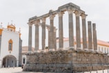 Ruins of an ancient Roman temple with multiple stone columns stand prominently, adjacent to a white building with orange accents, possibly a church. The temple's weathered stones contrast with the modern, well-maintained building beside it.