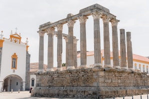 Ruins of an ancient Roman temple with multiple stone columns stand prominently, adjacent to a white building with orange accents, possibly a church. The temple's weathered stones contrast with the modern, well-maintained building beside it.