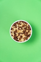 A white bowl filled with a mixture of chocolate and vanilla flavored ring-shaped cereal pieces sits against a bright green background.