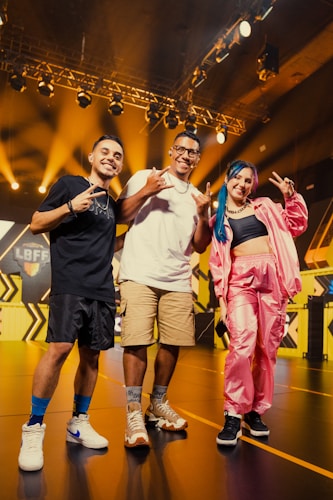 Three individuals are standing on a stage with bright stage lighting and a black and yellow geometric background. Two men and one woman are smiling and posing playfully with hand signs. The woman is dressed in a pink outfit with multicolored hair. The scene has a vibrant and energetic atmosphere.
