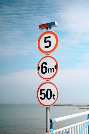 A series of circular traffic signs on a pole, each with different restrictions. The top sign shows a number 5, indicating a speed limit of 5 kilometers or miles per hour. The middle sign displays 6m, suggesting a height restriction of 6 meters. The bottom sign has 50t, representing a weight limit of 50 tons. The pole is topped with red and blue lights, possibly indicating it's near a regulated area. A metal fence is visible in the foreground with a view of the ocean and a mostly clear sky in the background.