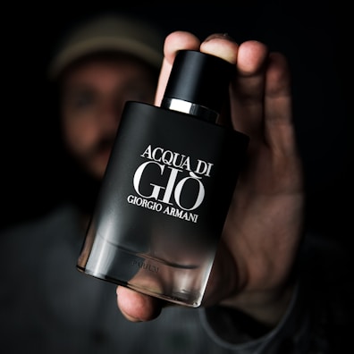 A hand holding a sleek, darkly designed Acqua di Giò perfume bottle by Giorgio Armani. The background is blurred, emphasizing the focus on the bottle.