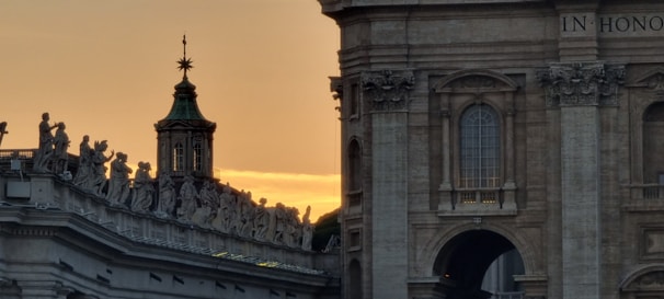 A detailed architectural scene featuring an ornate building with intricate sculptures adorning the roofline. The background is lit by a warm golden sunset, casting a serene glow on the stone surfaces.