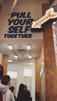A group of people stand in line inside a corridor with wooden walls and ceiling lights. Above them, a motivational sign reads 'PULL YOUR SELF TOGETHER' in bold letters, accompanied by an illuminated arrow pointing to the right.