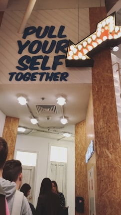A group of people stand in line inside a corridor with wooden walls and ceiling lights. Above them, a motivational sign reads 'PULL YOUR SELF TOGETHER' in bold letters, accompanied by an illuminated arrow pointing to the right.