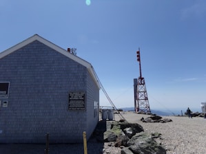 A building with gray siding stands in a rocky, open area beneath a clear blue sky. Signage on the building indicates it is an office and mentions a record high wind speed. Nearby, there is a tall metal structure with red markers. A person can be seen walking in the background.