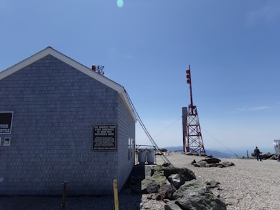 A building with gray siding stands in a rocky, open area beneath a clear blue sky. Signage on the building indicates it is an office and mentions a record high wind speed. Nearby, there is a tall metal structure with red markers. A person can be seen walking in the background.