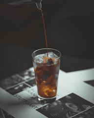 Coffee is being poured into a transparent glass filled with ice cubes, creating a swirling effect as it mixes with the contents. The glass is placed on a printed surface featuring images and text, with a darker background that contrasts with the lighter elements.