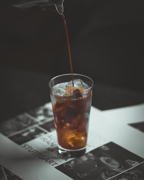 Coffee is being poured into a transparent glass filled with ice cubes, creating a swirling effect as it mixes with the contents. The glass is placed on a printed surface featuring images and text, with a darker background that contrasts with the lighter elements.